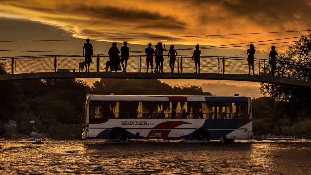 Puente peatonal con personas encima observando el cielo y un ómnibus pasando por debajo